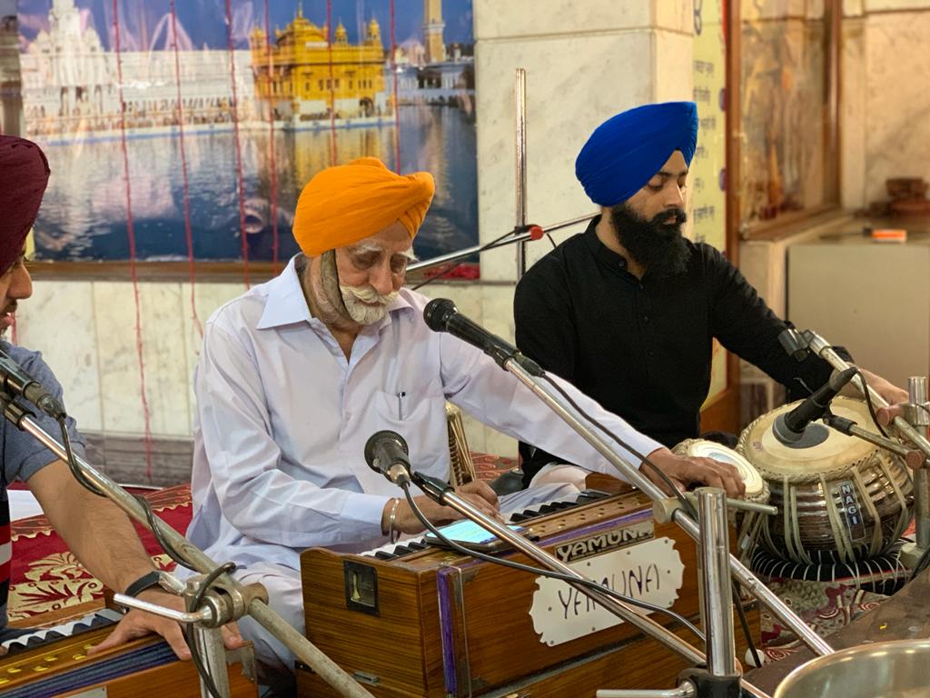 Musicians playing the harmonium and tabla during kirtan in front of a Golden Temple painting