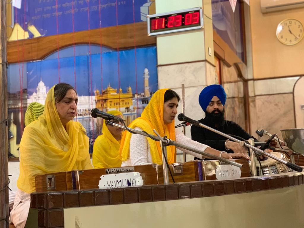 Women and a man performing kirtan at harmoniums in front of a Golden Temple mural